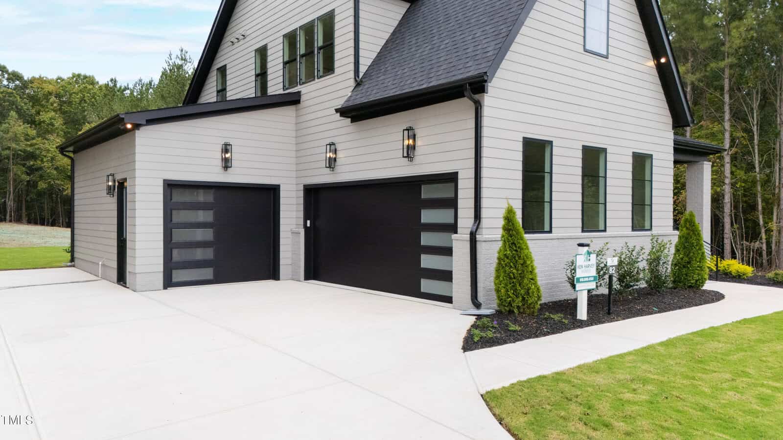 Modern house exterior with light gray siding, black trim, and two black garage doors with horizontal frosted glass panels. There is a clean concrete driveway, manicured lawn, shrubs, and a real estate sign near the entrance.