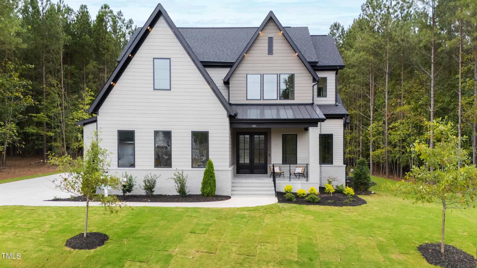 A modern two-story house with light-colored siding, black trim, and a covered front porch, surrounded by a freshly landscaped lawn and trees in the background.