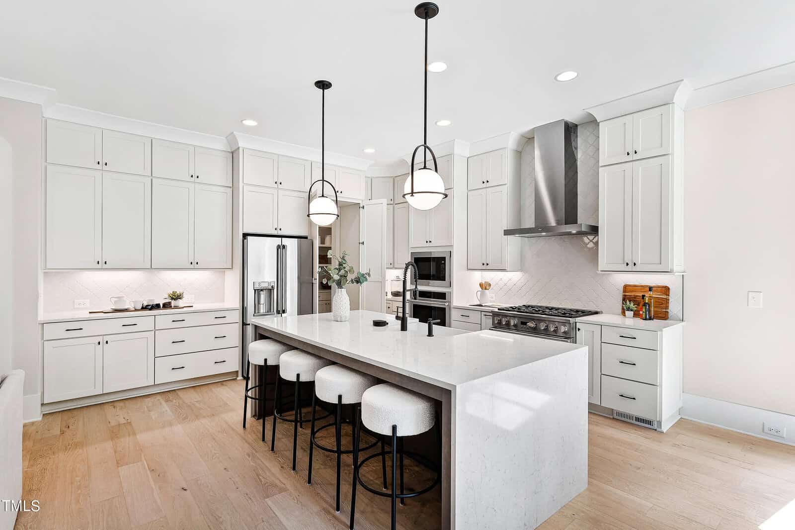 Modern white kitchen with shaker cabinets, stainless steel appliances, a large marble island with four cushioned stools, pendant lights above the island, and light wood flooring.