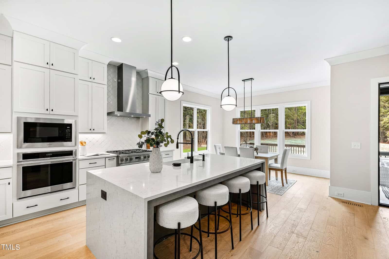 Modern kitchen with white cabinets, a marble island with four cushioned stools, stainless steel appliances, pendant lights, and a nearby dining area with large windows overlooking a wooded backyard.