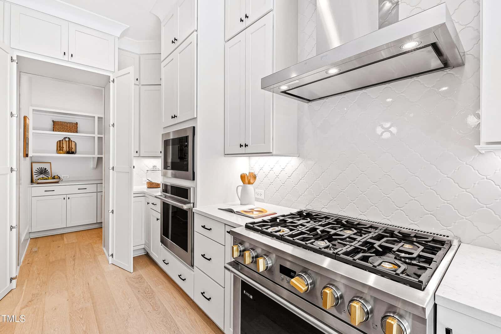 A modern, bright kitchen with white cabinets, a stainless steel gas stove and oven, a tiled backsplash, light wood flooring, and an adjacent walk-in pantry with open shelves and decorative items.