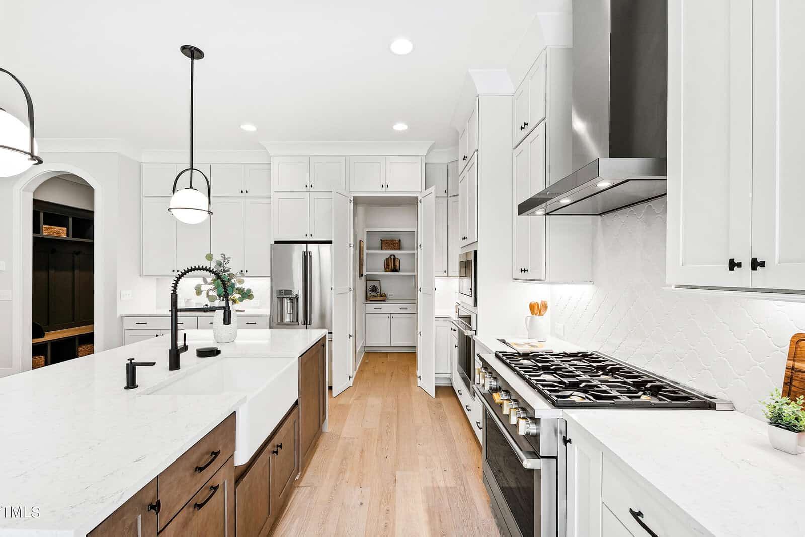 Bright, modern kitchen with white cabinets, stainless steel appliances, a large island with a farmhouse sink, gas stove, wood flooring, and a walk-in pantry with double doors in the background.
