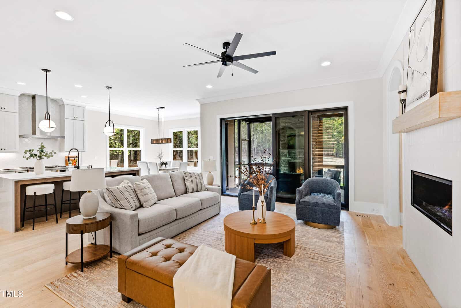 Modern open-concept living room with neutral decor, gray sofa, fireplace, round coffee table, and large sliding doors opening to a patio. Kitchen and dining area with pendant lights visible in the background.