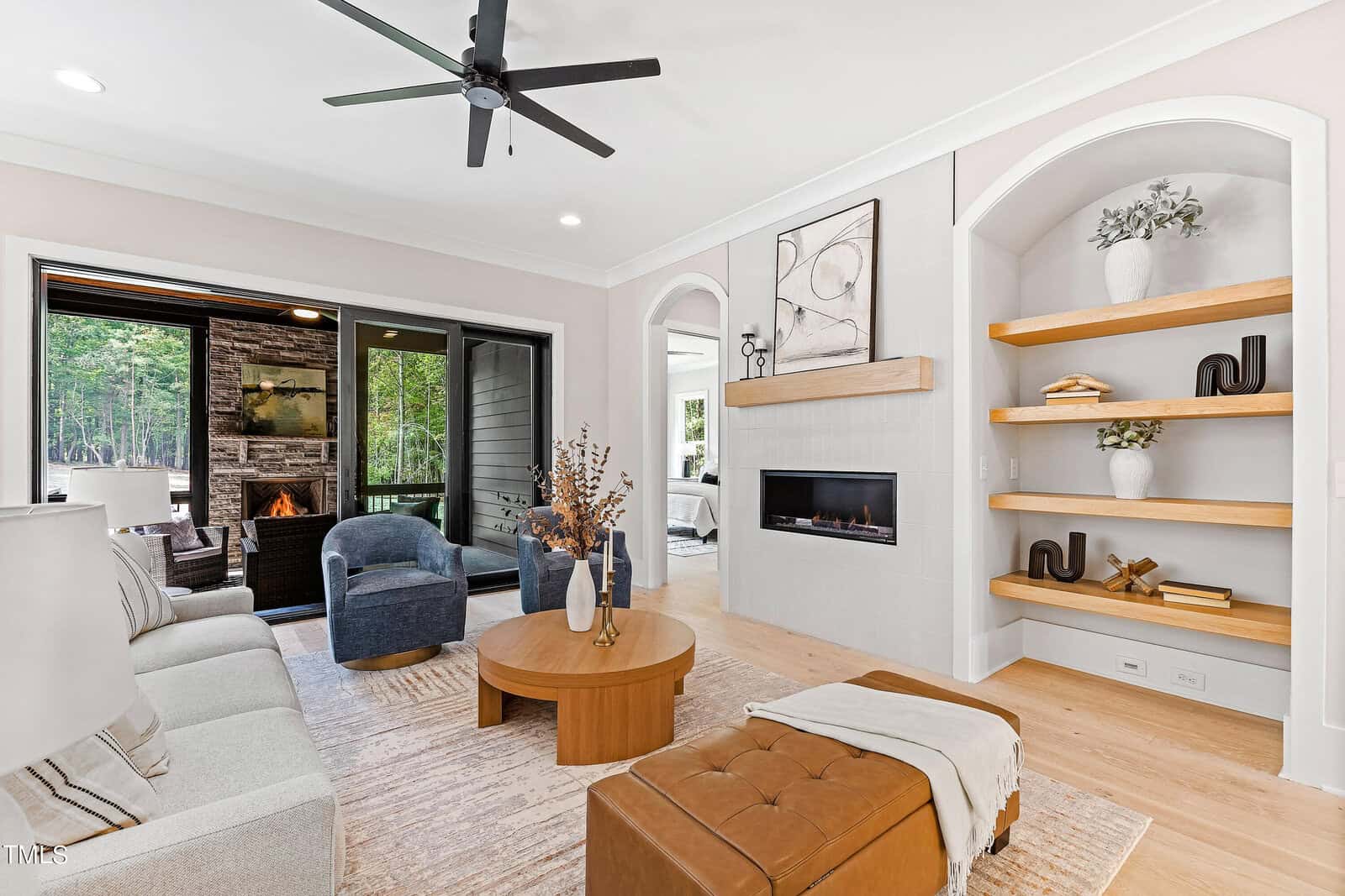 Modern living room with neutral decor, built-in shelves, round wooden coffee table, beige sofa, brown ottoman, and a fireplace. Large windows open to a patio with an outdoor fireplace and wooded view.