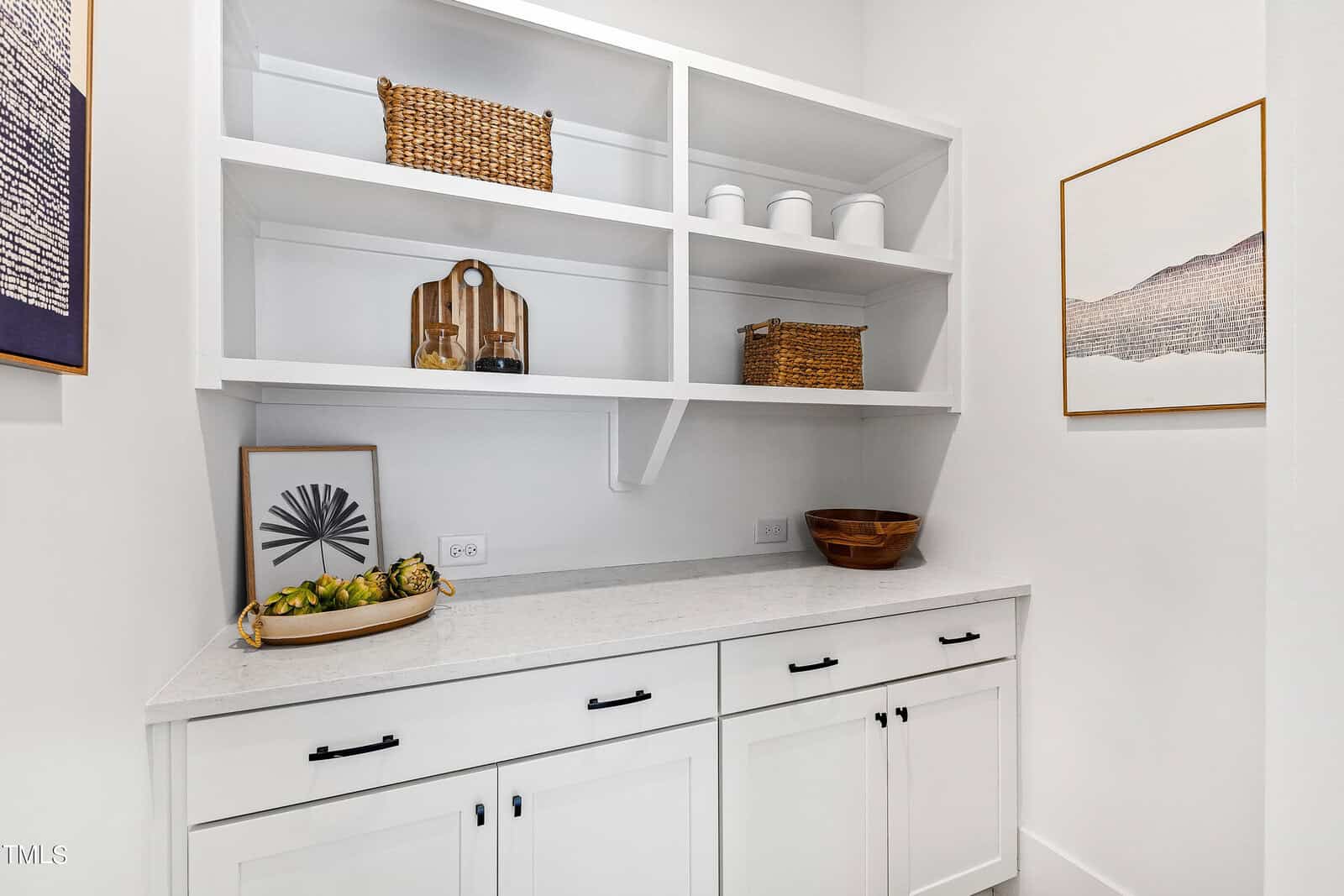 A small pantry with white cabinets and shelves, wicker baskets, white jars, a wooden bowl, cutting boards, and decorative artichokes. Minimalist decor and framed artwork on the right wall.
