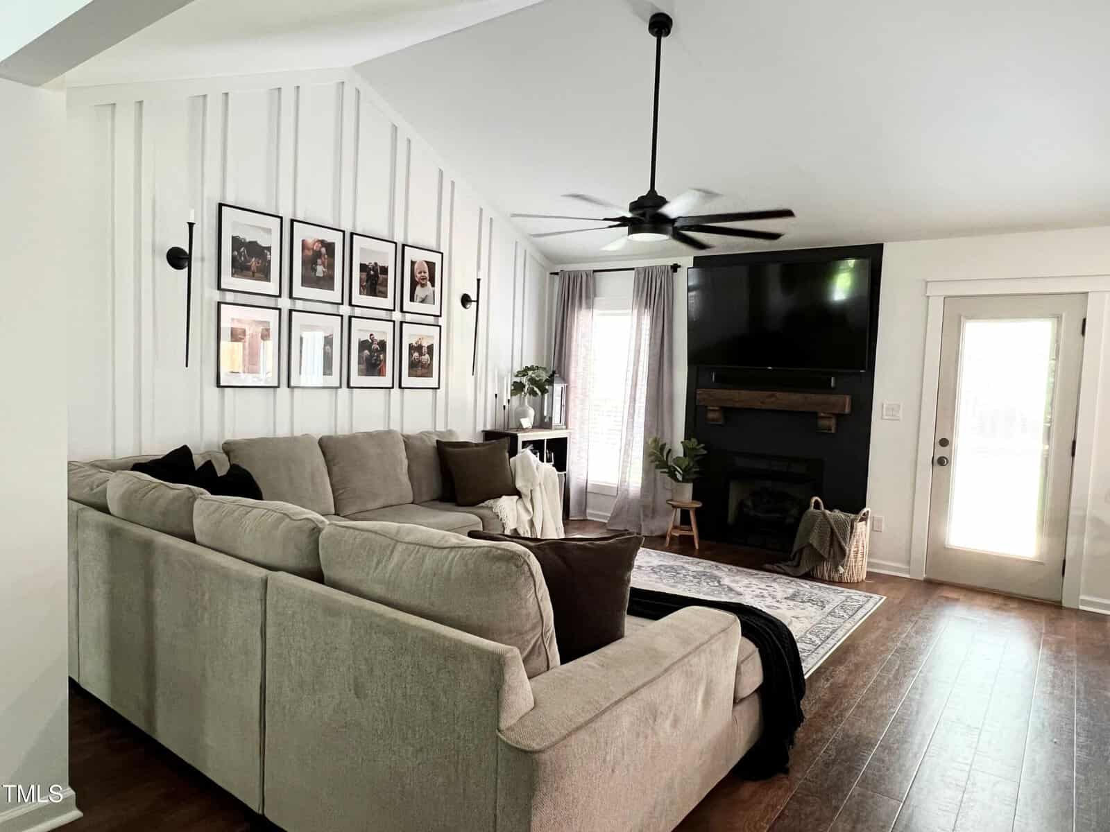 A cozy living room with a large beige sectional sofa, a wooden coffee table, and a patterned rug. The walls are decorated with framed photos. There's a fireplace with a mantel, a ceiling fan, and sheer curtains on the windows. A door leads outside.