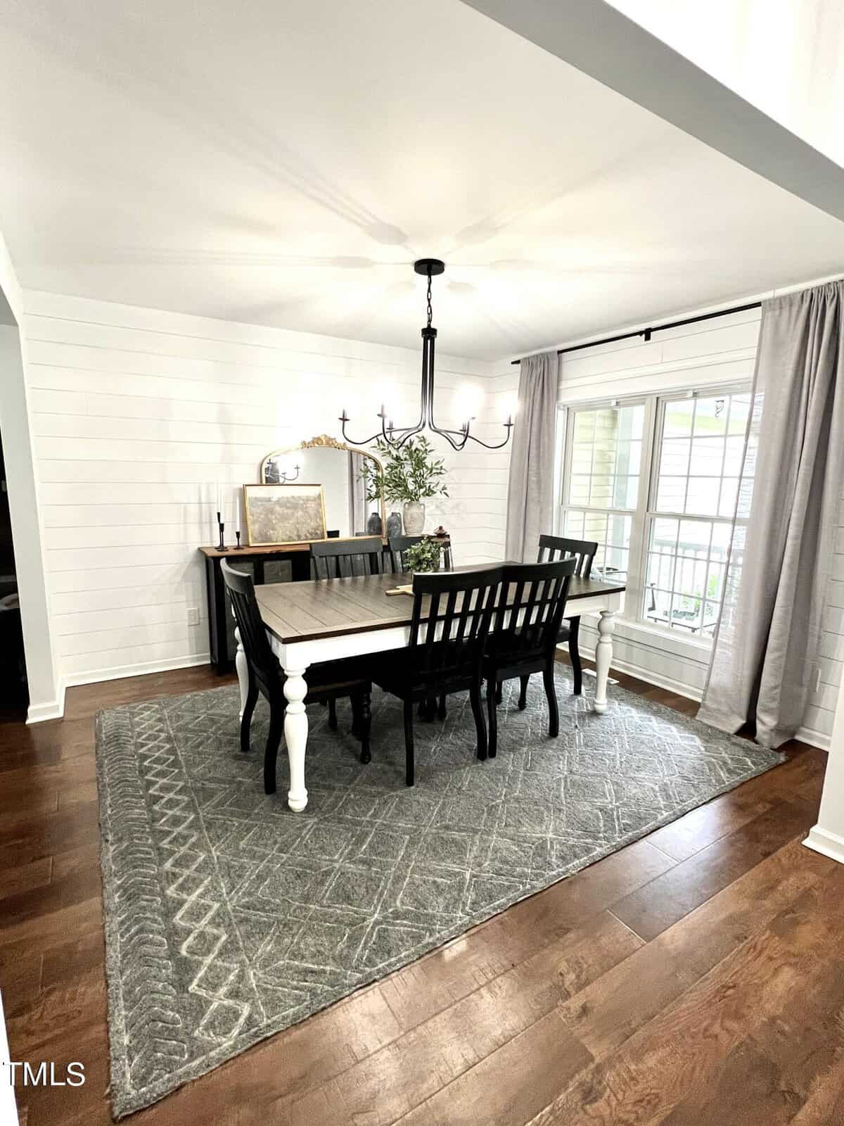 A dining room with a rectangular wooden table and six chairs. A chandelier hangs above. A side table holds decor and plants near a large window with light curtains. The room has white walls, hardwood floors, and a patterned gray rug.