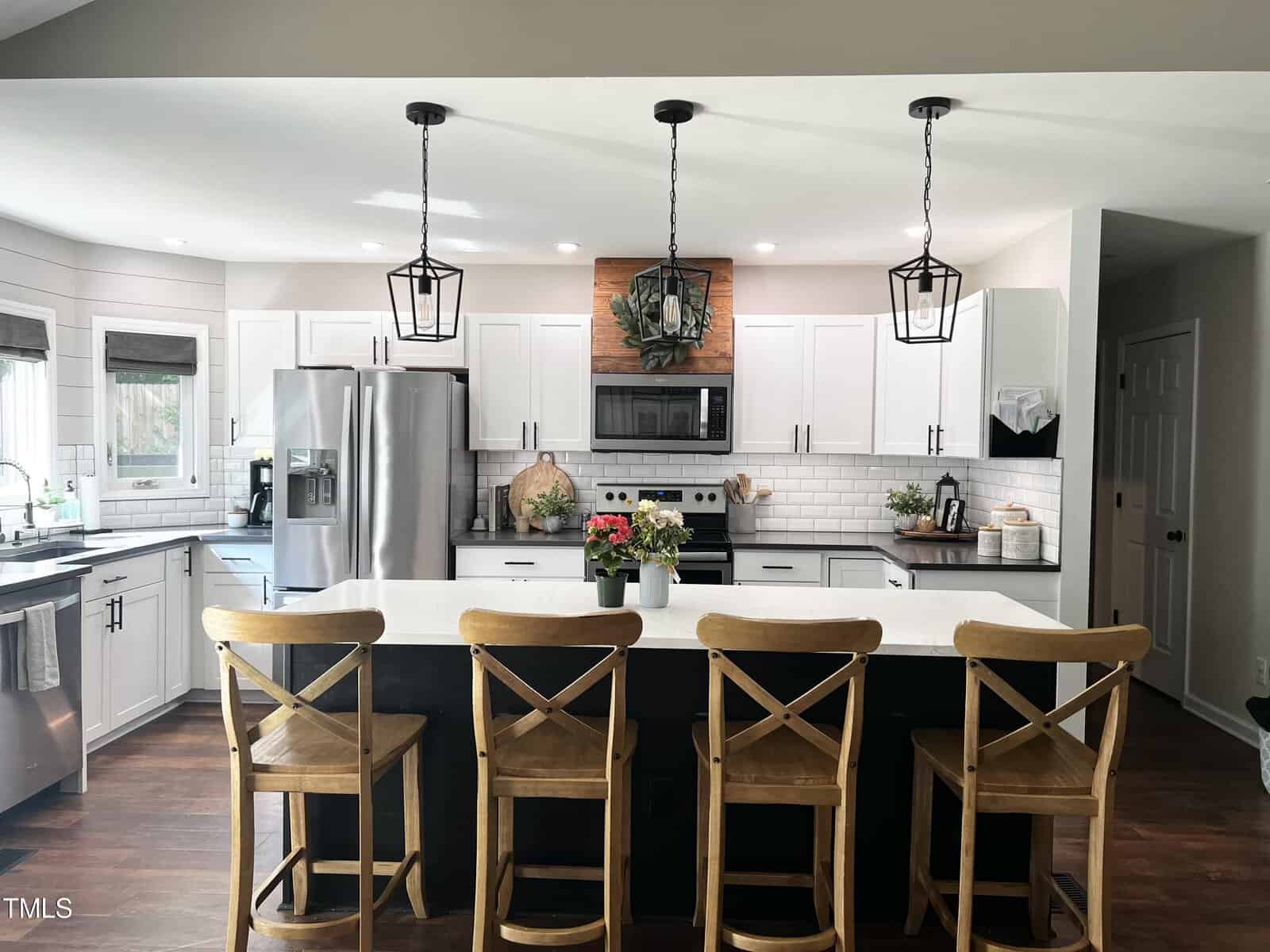 Modern kitchen with a large island featuring four wooden chairs. White cabinets and stainless steel appliances are present, along with pendant lights overhead. A decorative wreath is above the stove, adding a touch of green.