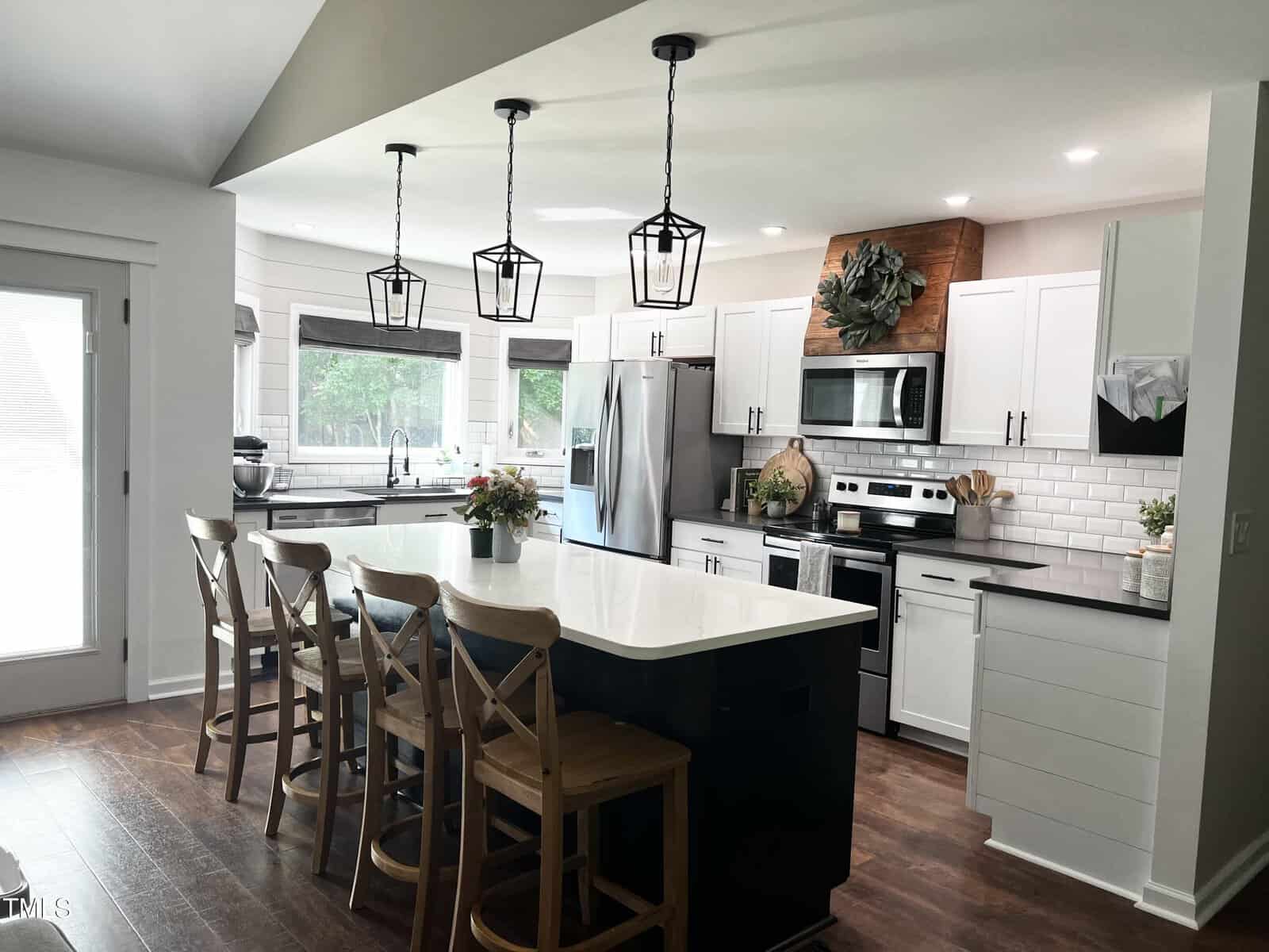 Modern kitchen with a large island, wooden stools, and pendant lights. Stainless steel appliances and white cabinets enhance the sleek design. A plant decorates the wall, and natural light fills the room through large windows.