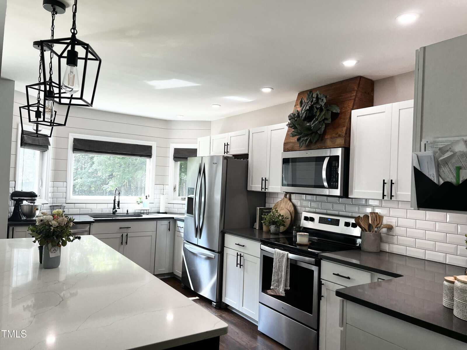 Modern kitchen with white cabinets and dark countertops. Stainless steel appliances include a fridge and an oven with a microwave above. A large island features hanging geometric light fixtures. A window allows natural light, and greenery decorates the space.