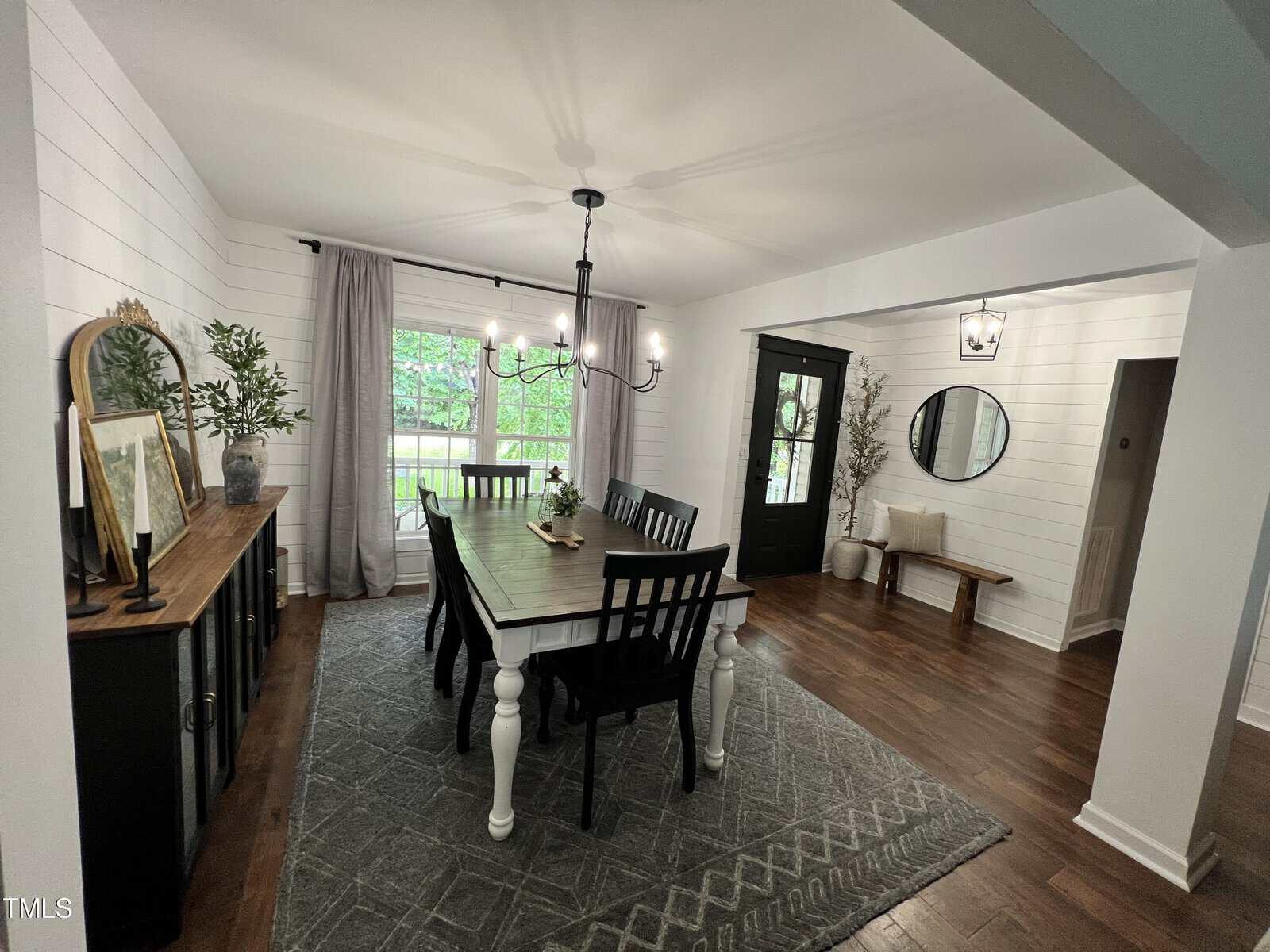 A dining room with a large white table, surrounded by black chairs. A decorative rug covers the wooden floor. A sideboard with artwork and plants is against the wall. A window with curtains lets in natural light. A door leads to another room.