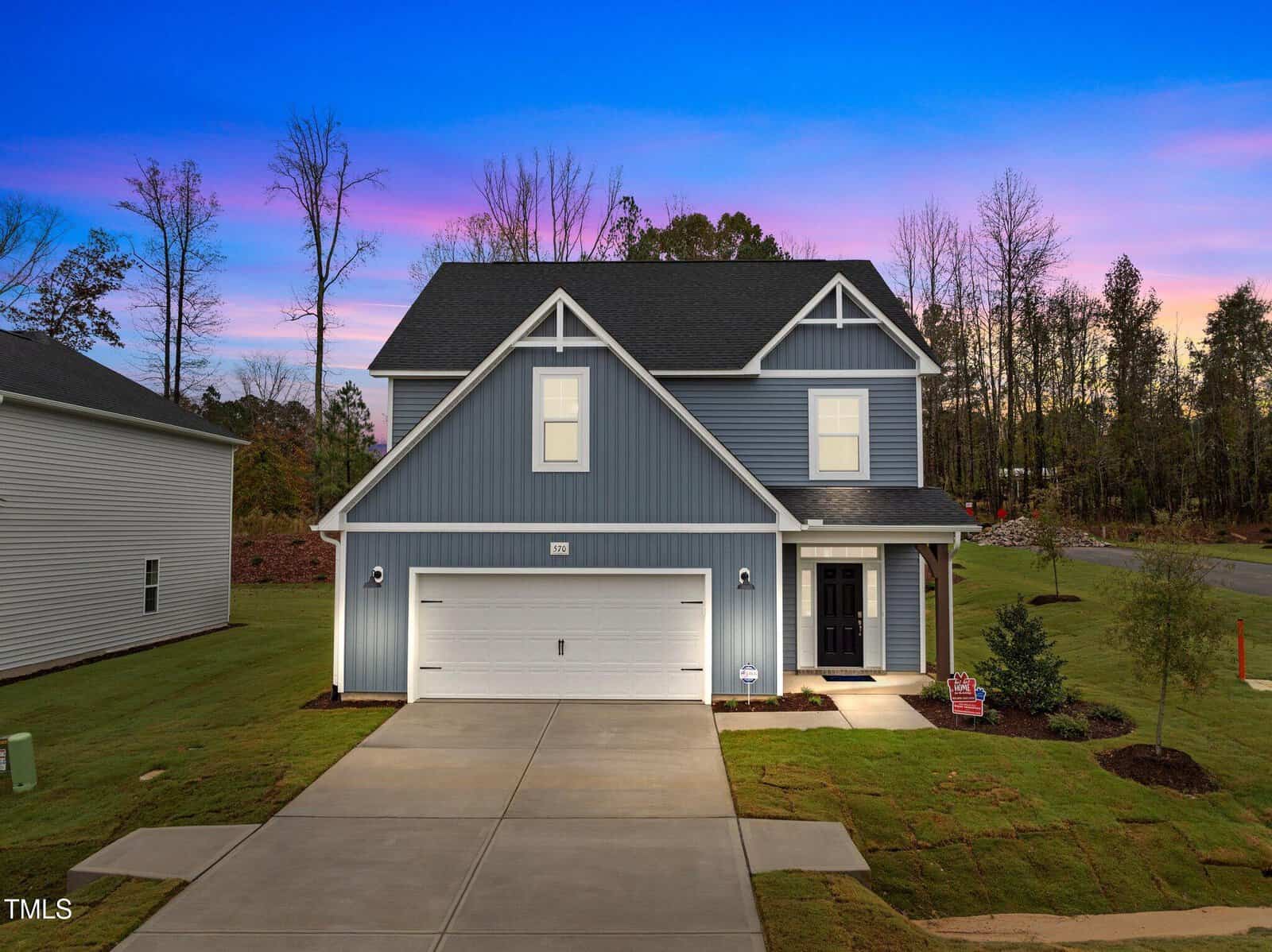 Two-story blue house with white trim, a double garage, and a covered entryway sits on a well-manicured lawn at sunset. The sky is pink and purple, and there are tall trees and a quiet street in the background.
