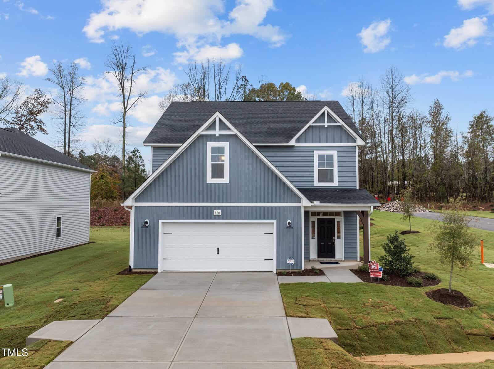 A two-story modern blue house with white trim, a double garage, and a covered front porch, set on a newly landscaped lawn with surrounding trees and a clear blue sky in the background.
