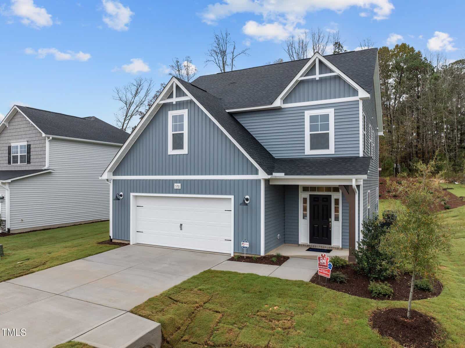 A modern two-story blue house with white trim, a double garage, black front door, and a well-kept lawn. The home sits on a concrete driveway, with trees and greenery in the background.