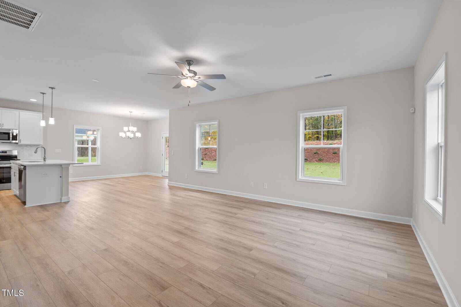 Spacious open-concept living area with light wood flooring, large windows, ceiling fan, and view of a modern kitchen with white cabinets and stainless steel appliances in the background.