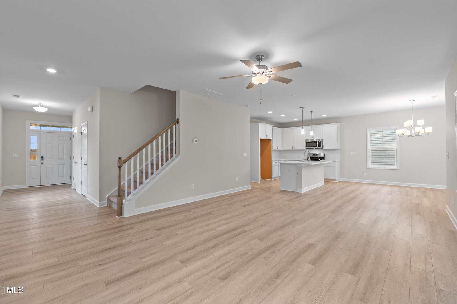 Spacious open-plan living area with light wood floors, neutral walls, ceiling fans, a staircase, and a modern kitchen with white cabinetry and stainless steel appliances. A chandelier hangs over the dining area.
