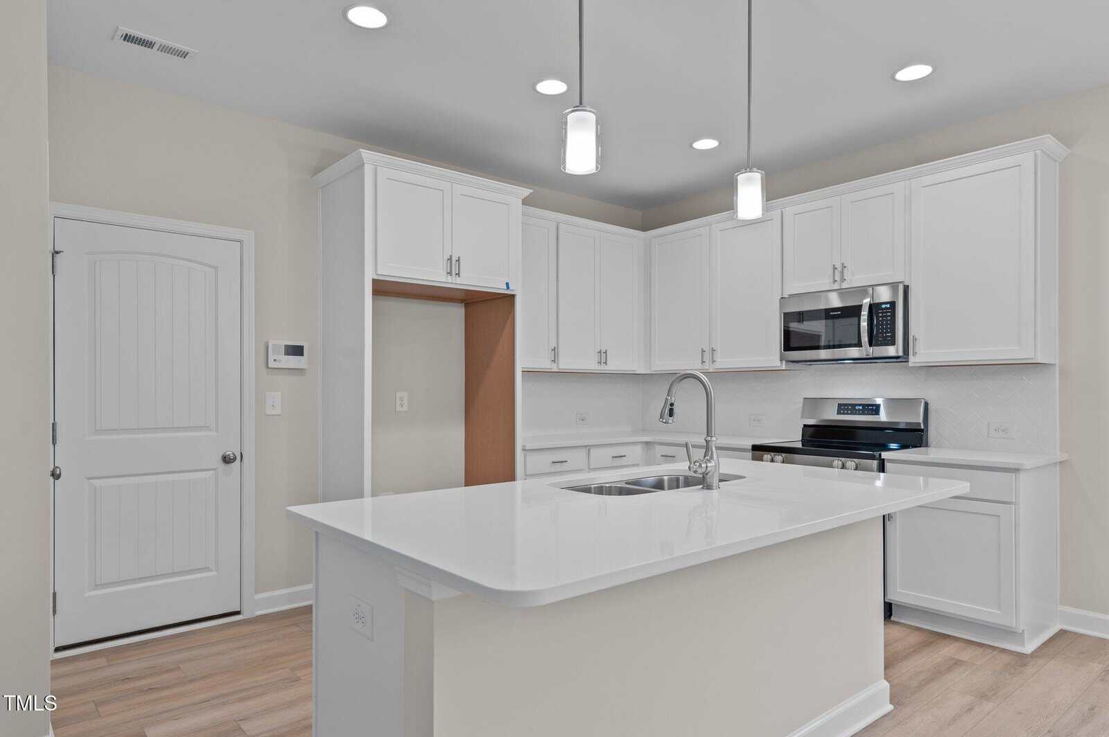 Modern kitchen with white cabinets, stainless steel appliances, a built-in microwave, and a large island with a sink. Light wood flooring and recessed lighting create a bright, clean, and open atmosphere.