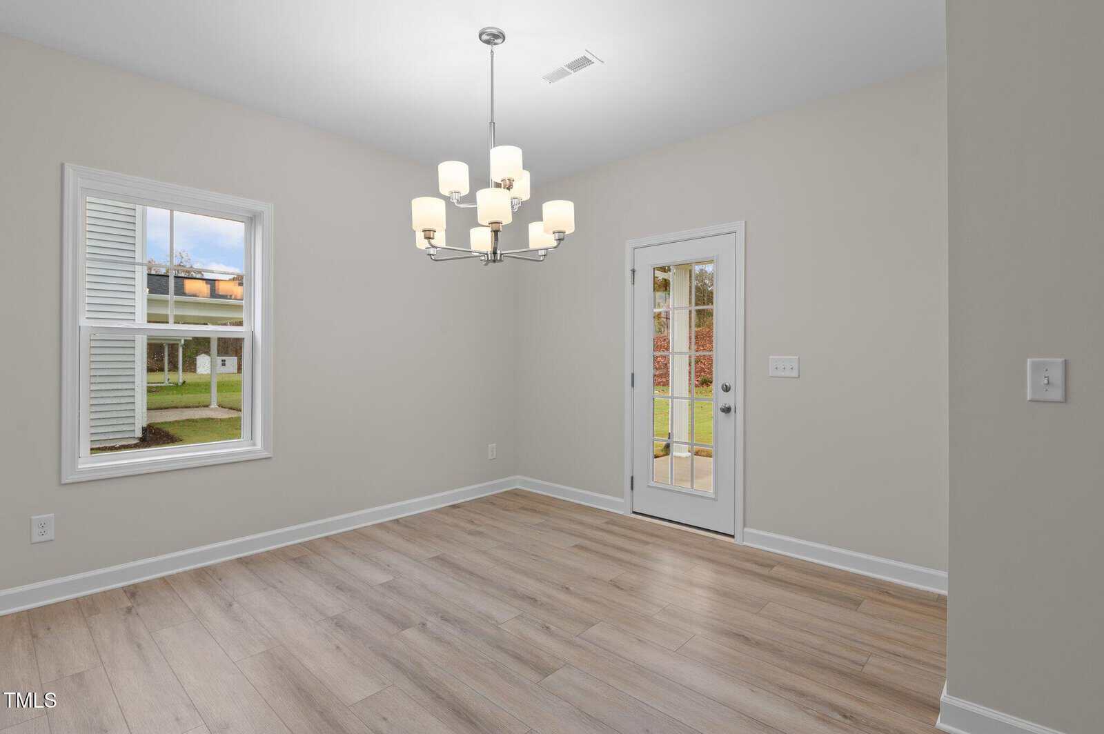 A bright, empty room with light wood flooring, beige walls, a modern chandelier, a window showing a yard, and a glass door leading outside. Natural light fills the space.