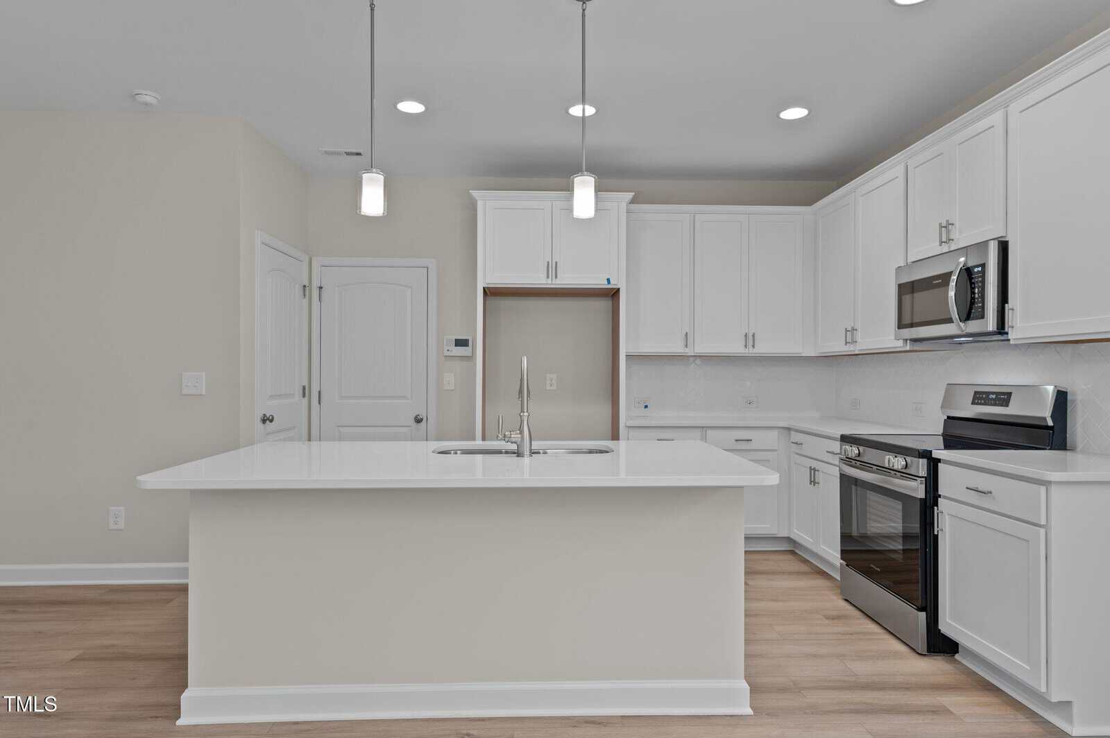 Modern kitchen with white cabinets, stainless steel appliances, a center island with a sink, pendant lighting, light wood flooring, and a space for a refrigerator. Walls and backsplash are also white, creating a bright, clean look.