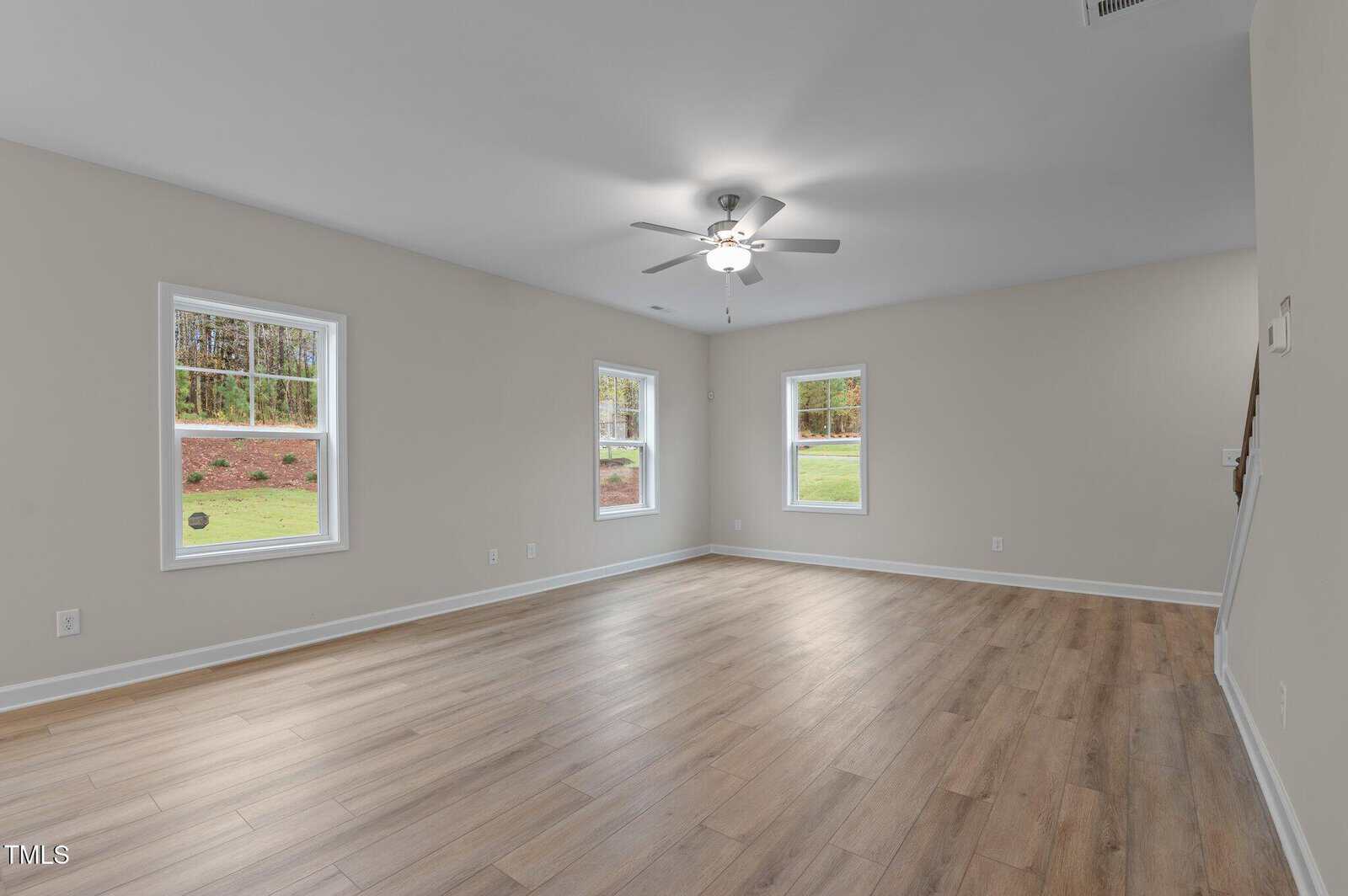 A spacious, empty room with light wood flooring, beige walls, three windows showing a grassy yard, and a ceiling fan with a light. Stairs are visible in the background on the right side.