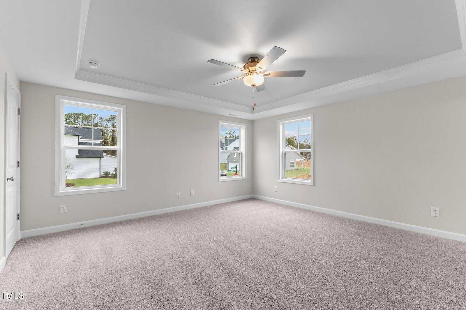 Spacious empty bedroom with beige walls, light gray carpet, three windows, a ceiling fan with a light, and white trim. Natural light fills the room through the windows.