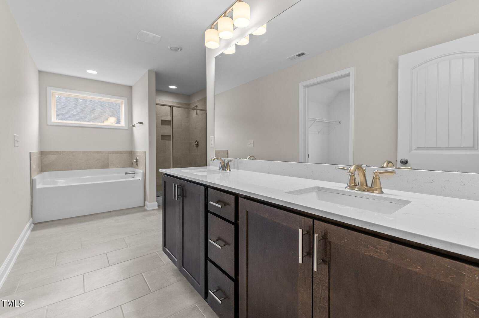 Modern bathroom with double sink vanity, large mirror, gold fixtures, soaking tub, walk-in shower, light gray tile floor, and neutral walls. A white door and window provide additional light and access.