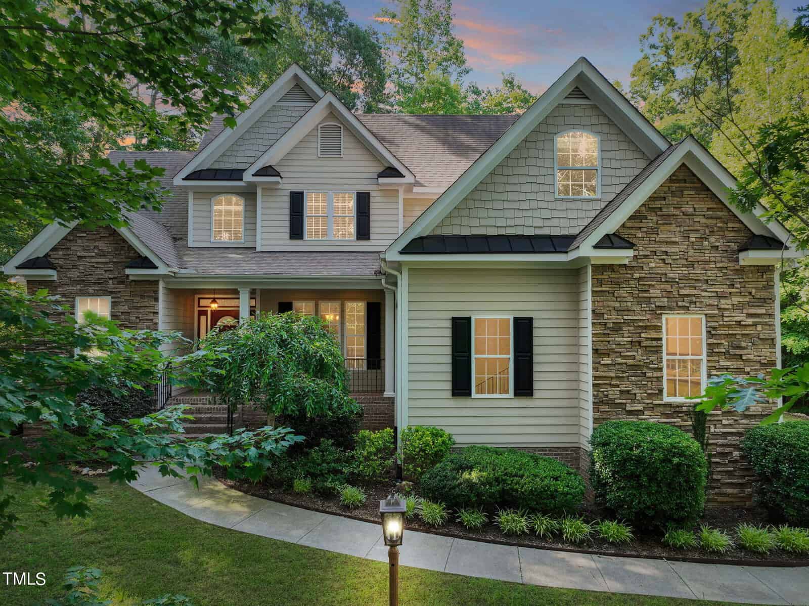 Nestled among trees in Wake Forest, this charming two-story house on 3585 Boulder Court features a blend of stone and siding exterior. With its peaked roof, shuttered windows, and welcoming front porch, a lit pathway guides you through the garden to the inviting entrance.