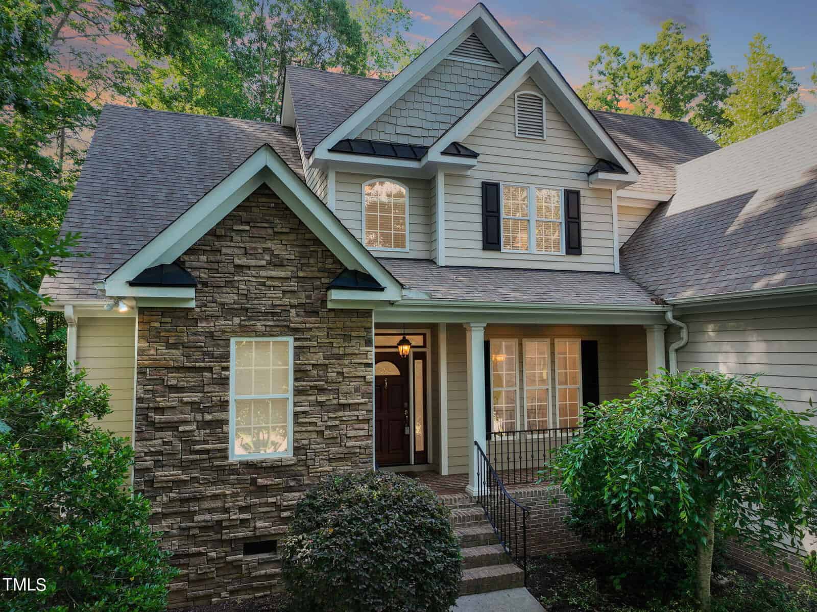 A charming two-story house with a stone and siding exterior, surrounded by lush greenery. It features a welcoming front porch and multiple windows. The roof is steeply pitched, and the sky is cloudy with hints of sunset colors.