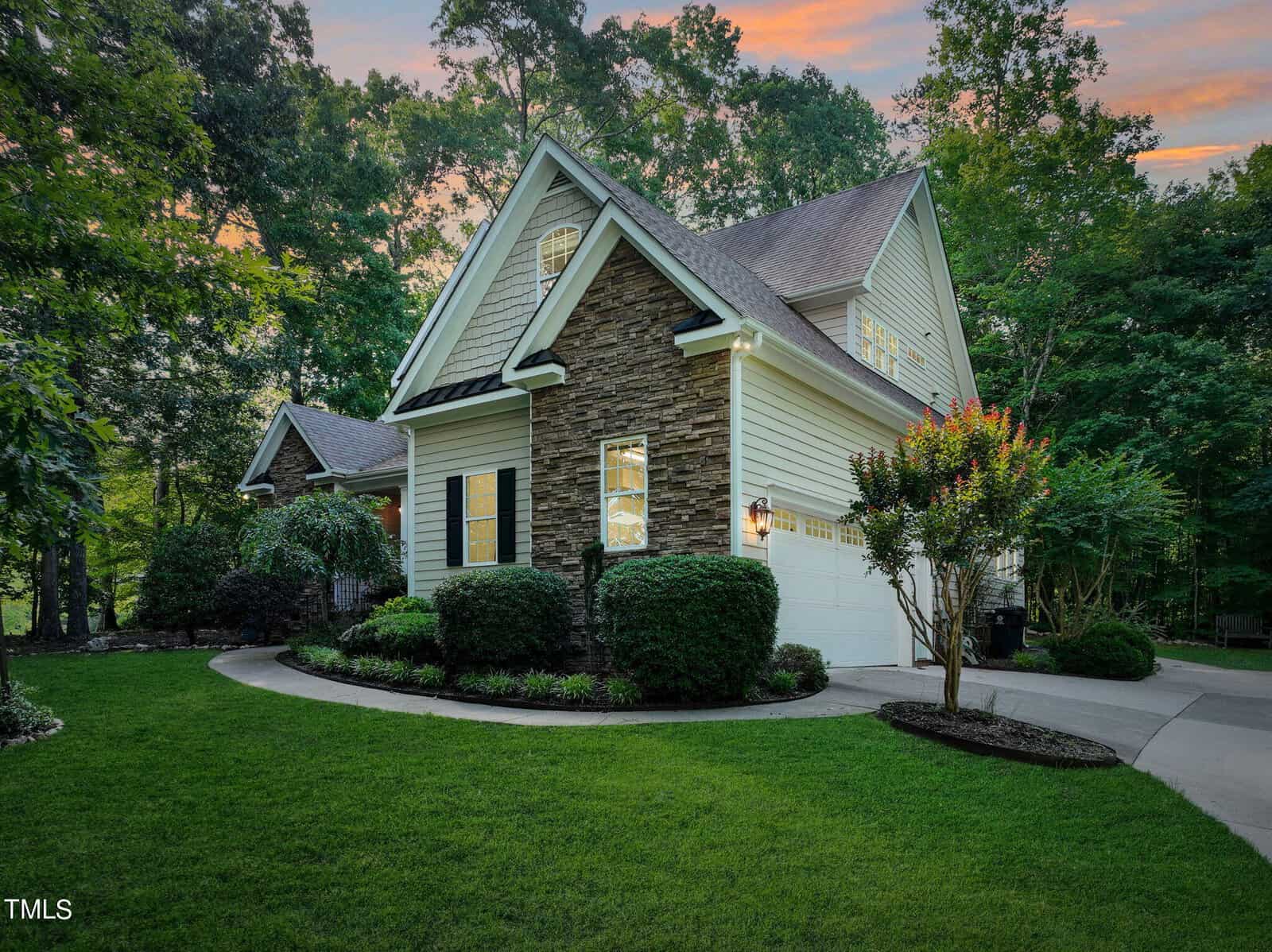 A charming house with stone accents and white siding is nestled among lush trees. It features a manicured lawn, bushes, and a double garage. The sky is a soft gradient of evening hues, adding warmth to the scene.