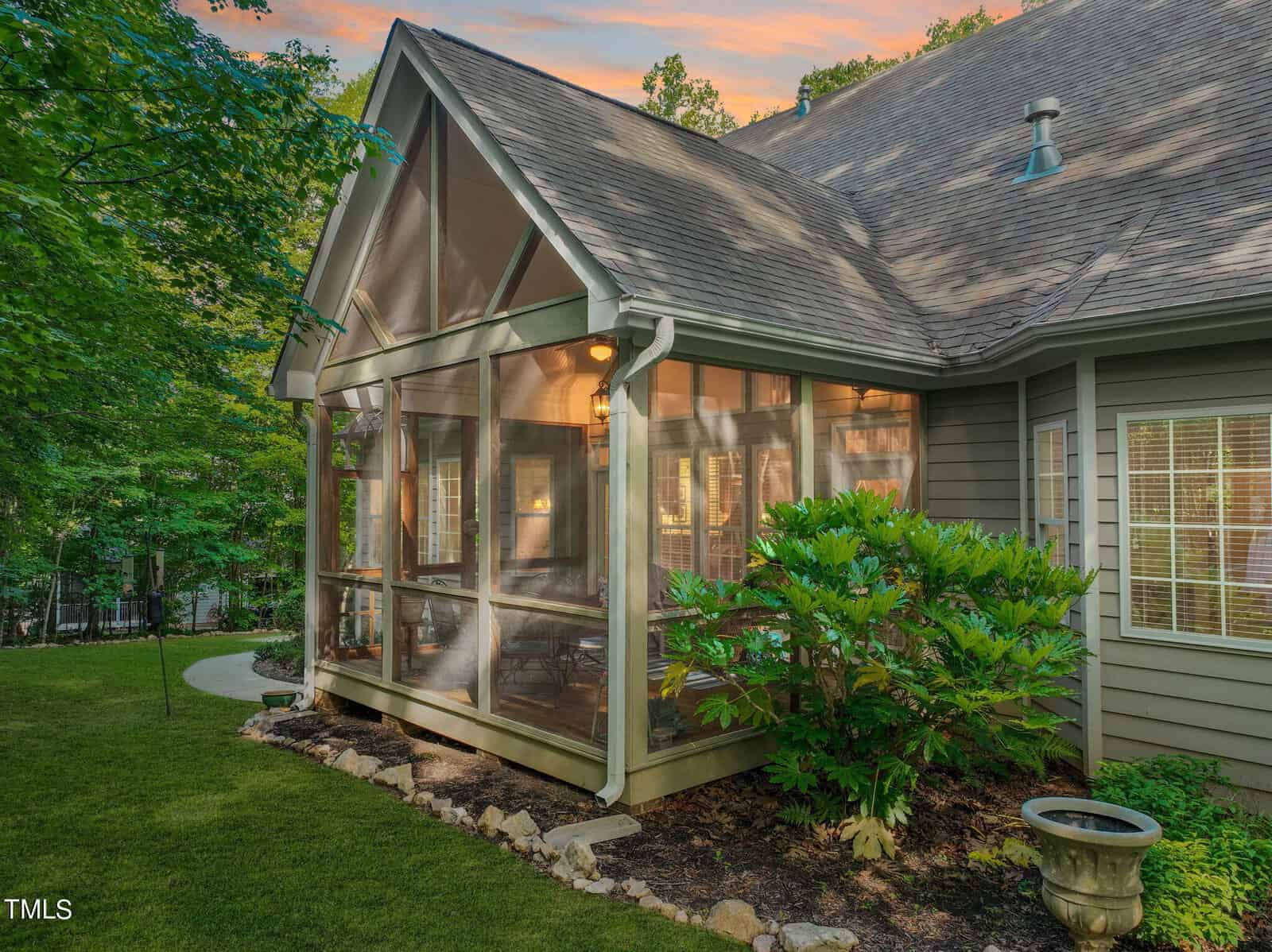 A screened porch with a peaked roof attached to a house, surrounded by greenery. Warm light glows from inside, and a pathway leads to the porch. The sky shows a colorful sunset.