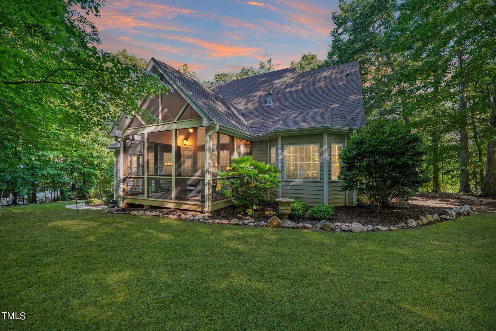 A charming house with a screened porch is nestled amid lush green trees. The manicured lawn extends around the home. The sky is a striking mix of orange and blue hues as the sun sets in the background.
