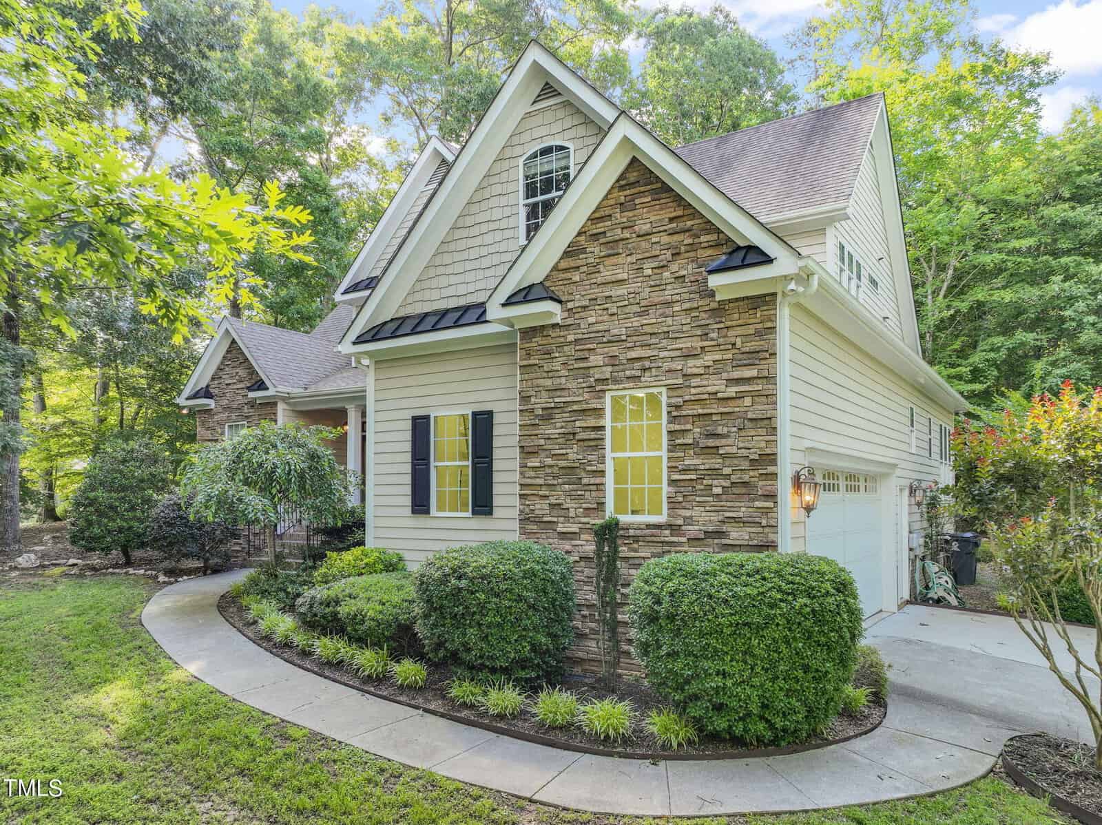 A charming two-story house with stone and siding exterior, surrounded by lush greenery. The home has a multi-gabled roof, shuttered windows, and a well-maintained front yard with shrubs and a curved concrete pathway leading to the entrance.