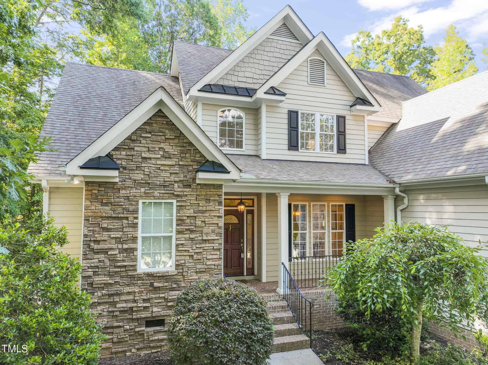 A two-story house with a stone facade and beige siding is surrounded by greenery. The front door, framed by windows with shutters, is accessible via a small stairway with railings. The roof is sloped with gables.