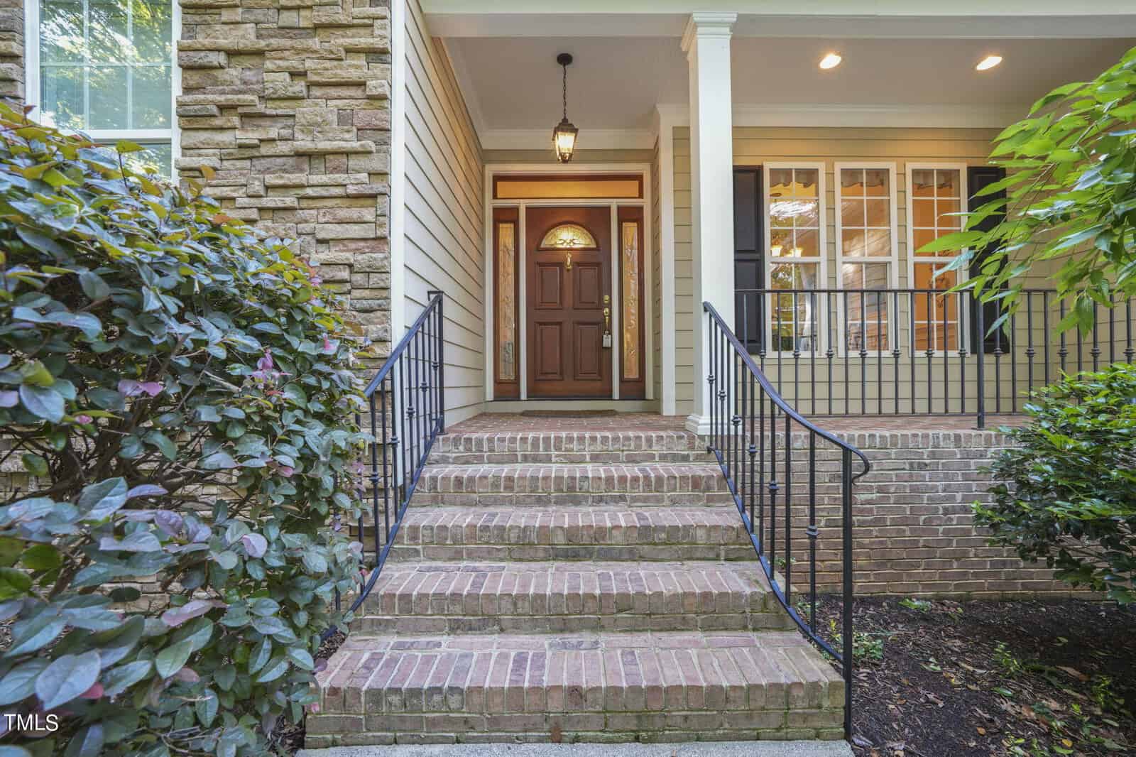 Front entrance of a house with a brick staircase leading to a wooden door. The entry is covered, with a hanging lantern and white pillars. Shrubs border both sides of the steps, and a window with white shutters is visible on the right.