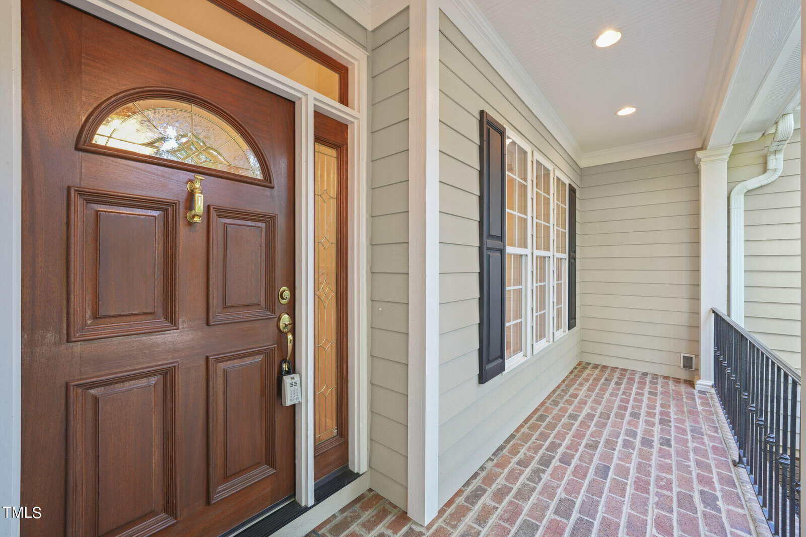 A polished wooden front door with a decorative glass panel and brass knocker on a house with beige siding. The entryway is bordered by a brick floor and features a railing on the right side and large windows with shutters on the left.