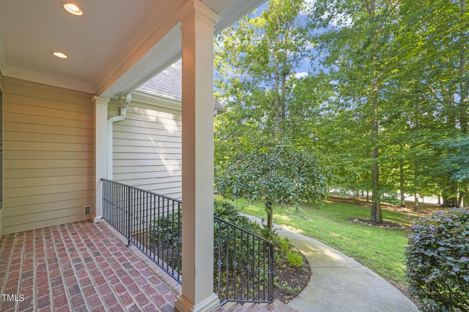 A porch with brick flooring and beige siding overlooks a lush green garden. A metal railing borders the edge, and a concrete path curves through the grass, surrounded by tall trees under a clear blue sky.