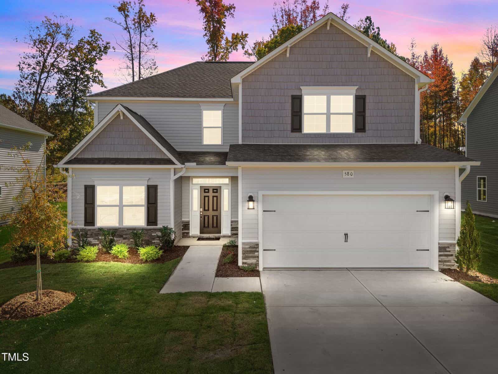 A two-story suburban house with gray siding, a double garage, a well-manicured lawn, and illuminated windows and exterior lights, set against a sunset sky with trees in the background.