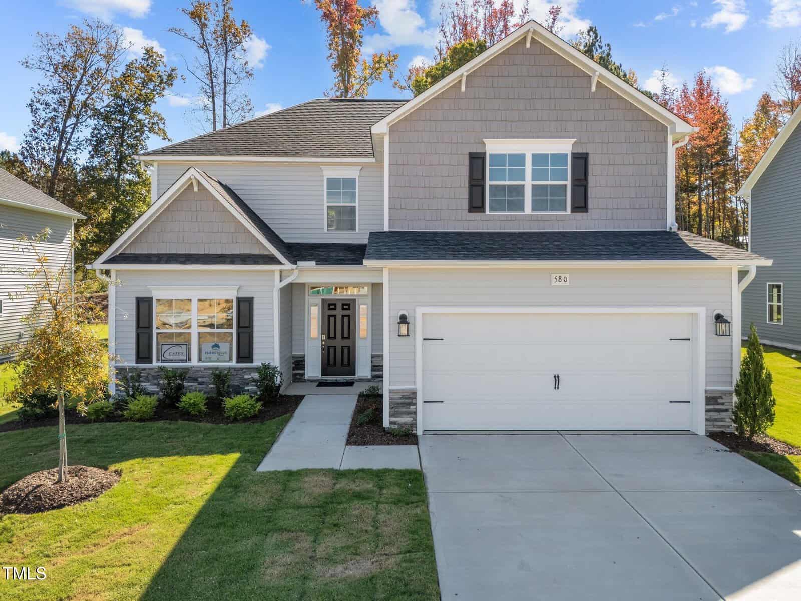 Two-story suburban house with gray siding, white trim, black shutters, and a double garage. A manicured lawn, small shrubs, and a young tree decorate the front yard under a blue sky with scattered clouds.