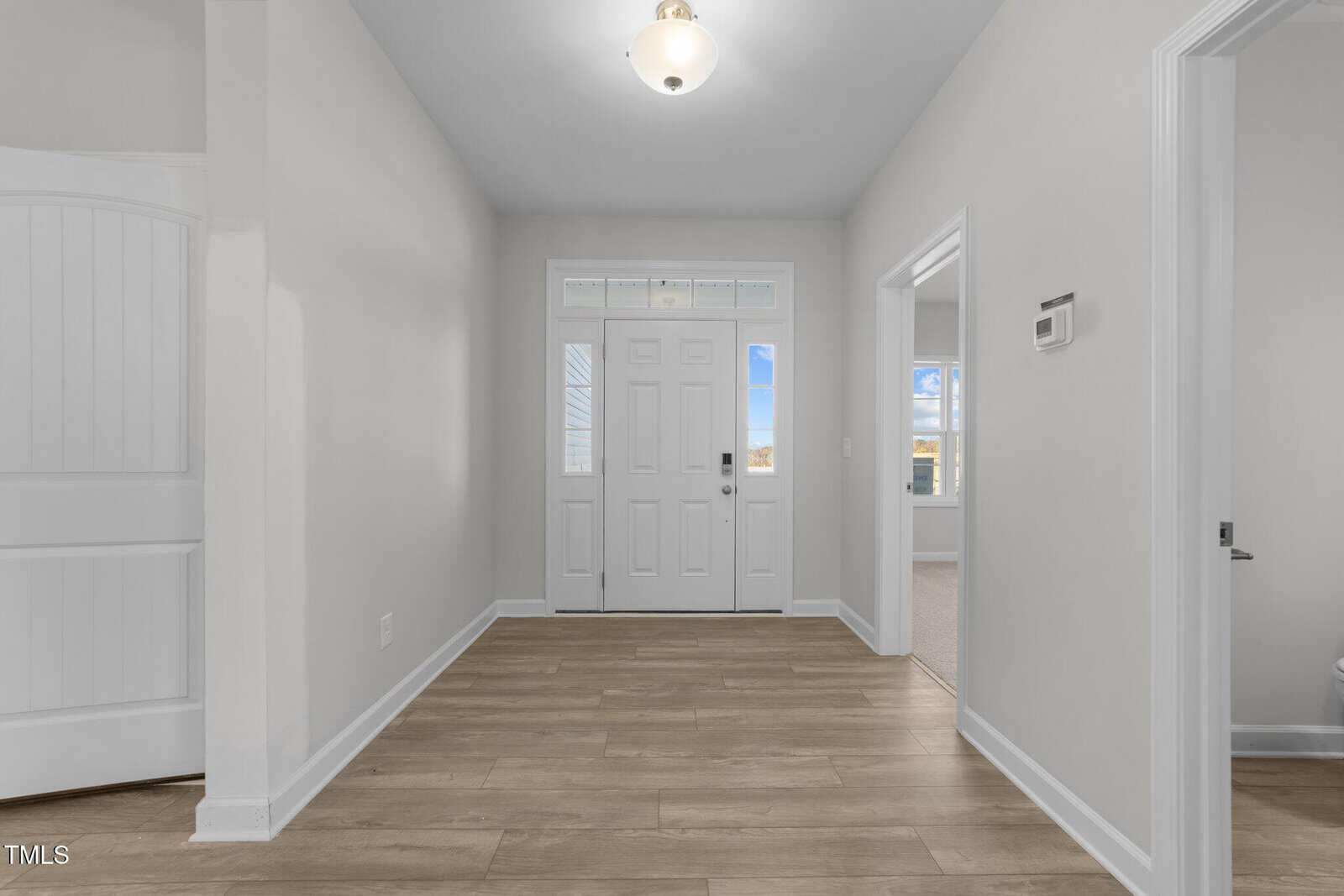 A bright entryway with light-colored walls, wood flooring, a white front door with glass panels, and an overhead light fixture. Two open doorways lead to other rooms, and a thermostat is mounted on the right wall.