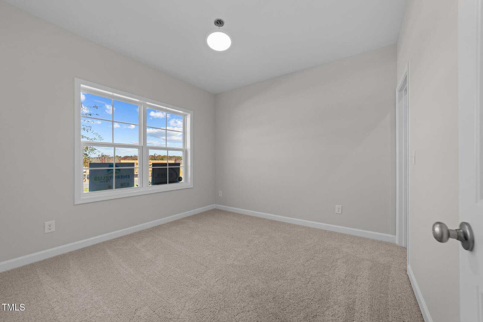 A small, empty room with beige carpet, light gray walls, a white ceiling, a round ceiling light, and a window letting in natural light with a view of trees and a blue sky outside.