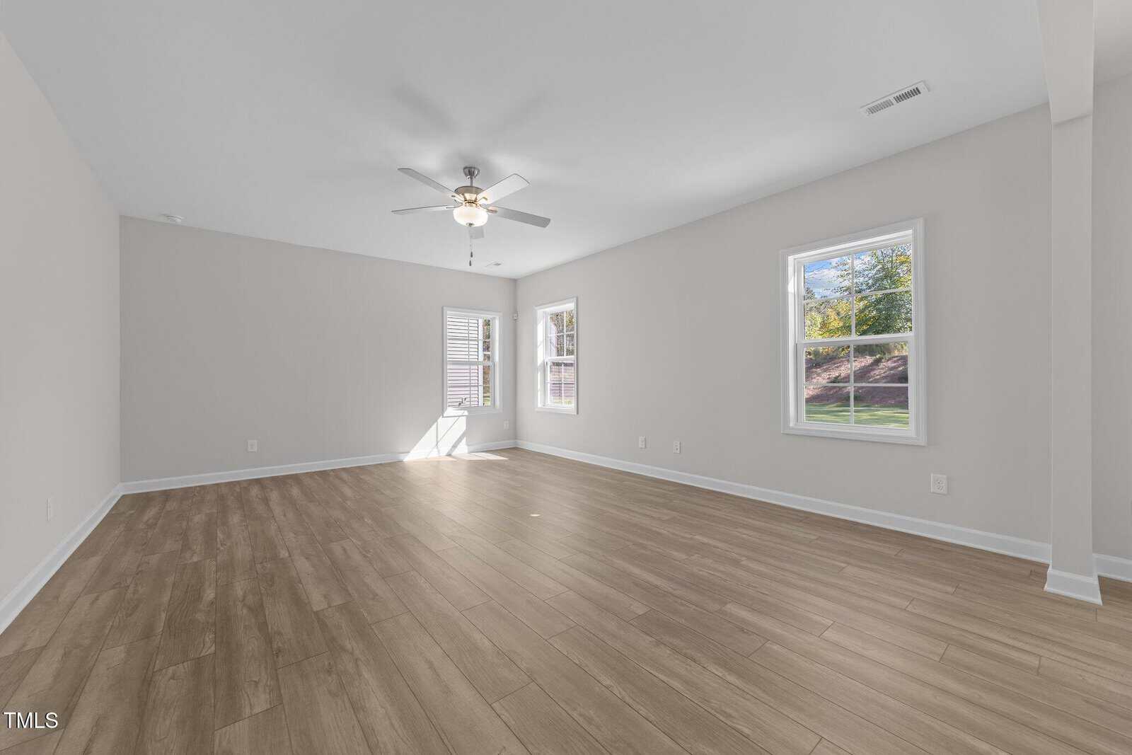 A spacious, empty room with light-colored walls, wood-style flooring, two windows letting in natural light, and a ceiling fan with a light fixture.