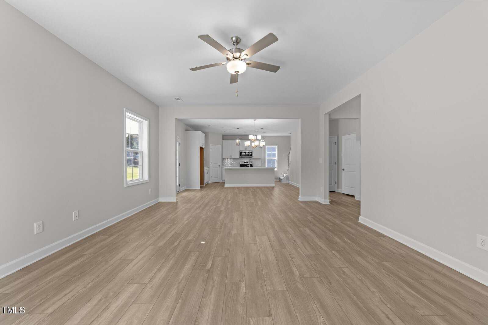Spacious, empty living area with light wood flooring, white walls, ceiling fan, and view into a modern kitchen with white cabinets and pendant lights. Natural light enters through windows on the left.
