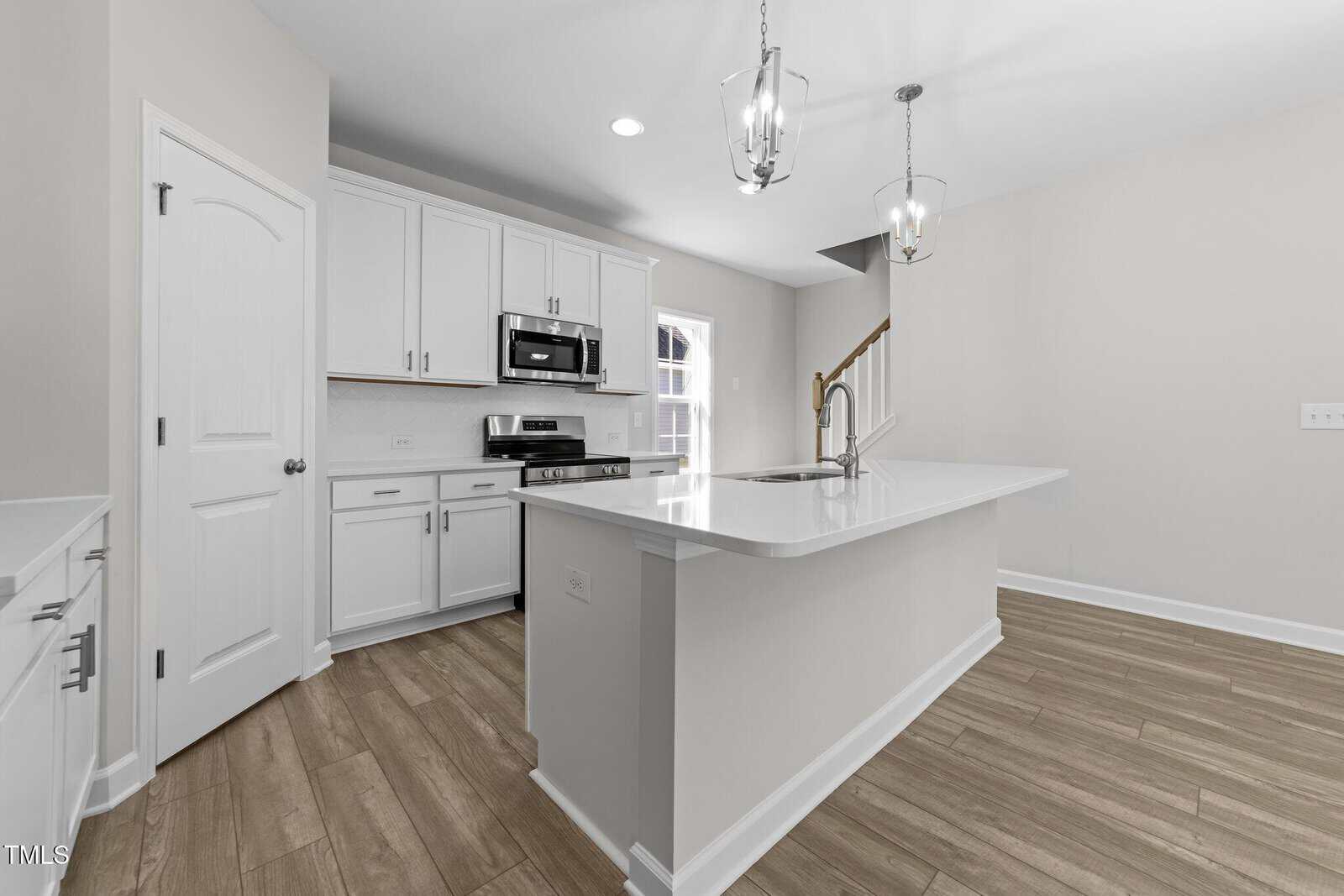 Modern kitchen with white cabinets, stainless steel appliances, a large island with sink, pendant lighting, light-colored walls, and wood-look flooring. A door and staircase are visible in the background.