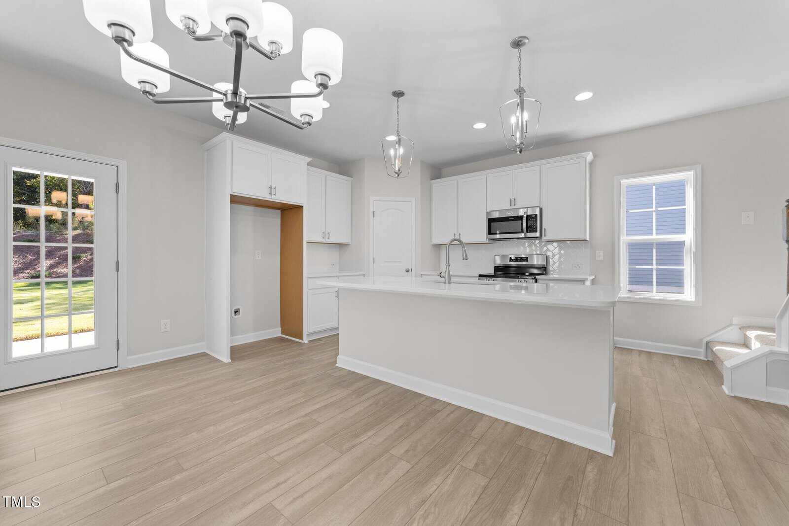 Modern kitchen with white cabinets, stainless steel appliances, large island with sink, and light wood flooring. Brightly lit by natural light from a window and glass door. Contemporary light fixtures hang from the ceiling.
