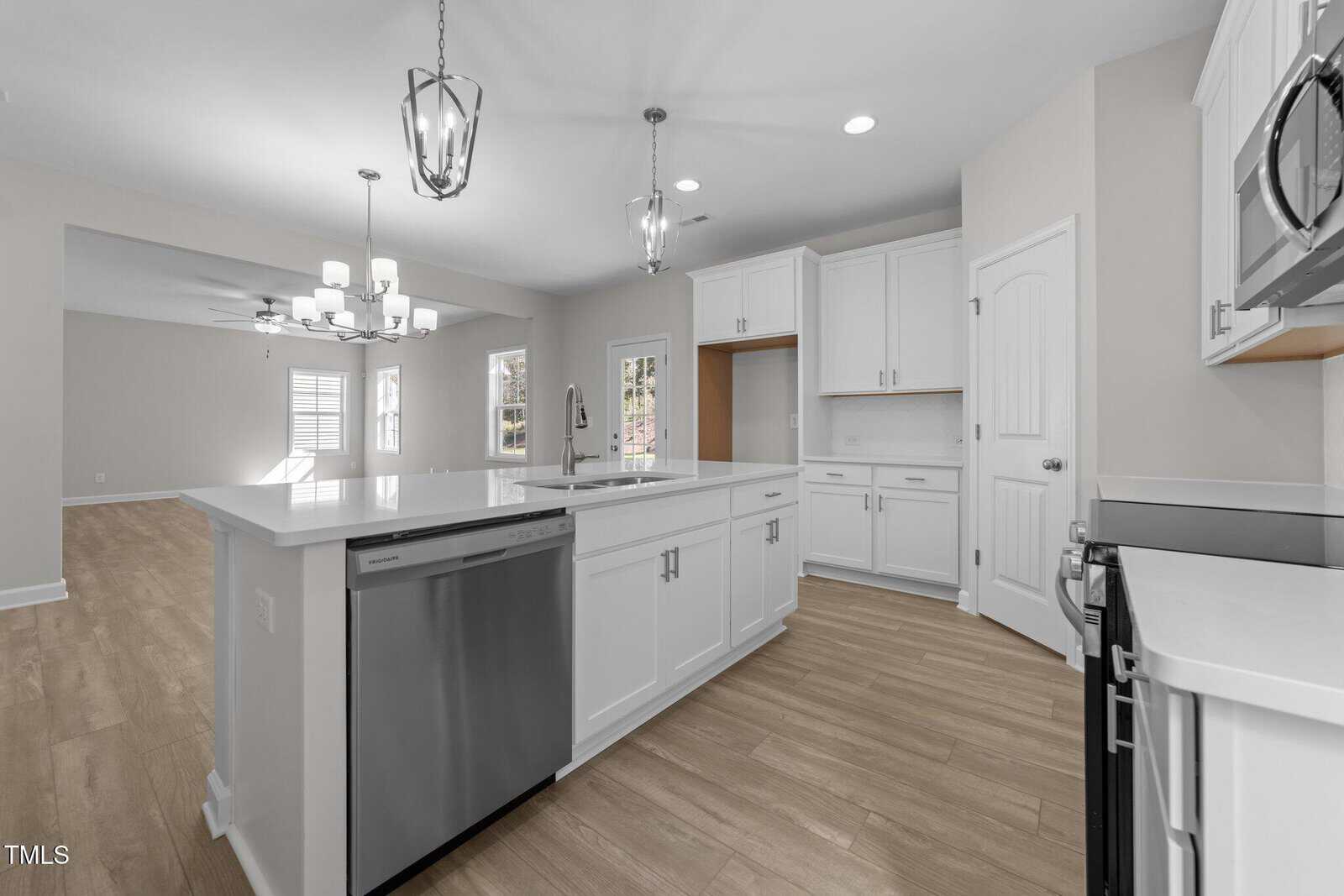 Bright modern kitchen with white cabinets, stainless steel appliances, pendant lights over a large island, and light wood flooring; open layout leads to a spacious living area with windows and a ceiling fan.