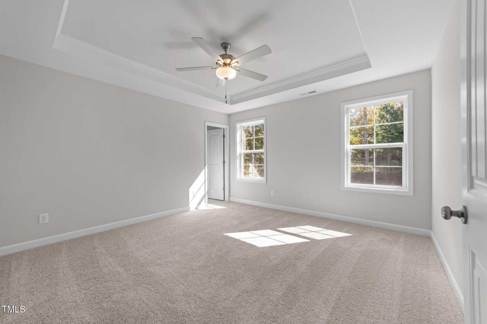 Bright, empty bedroom with beige carpet, light gray walls, tray ceiling, ceiling fan, two large windows letting in sunlight, and a white door leading to an adjacent room.