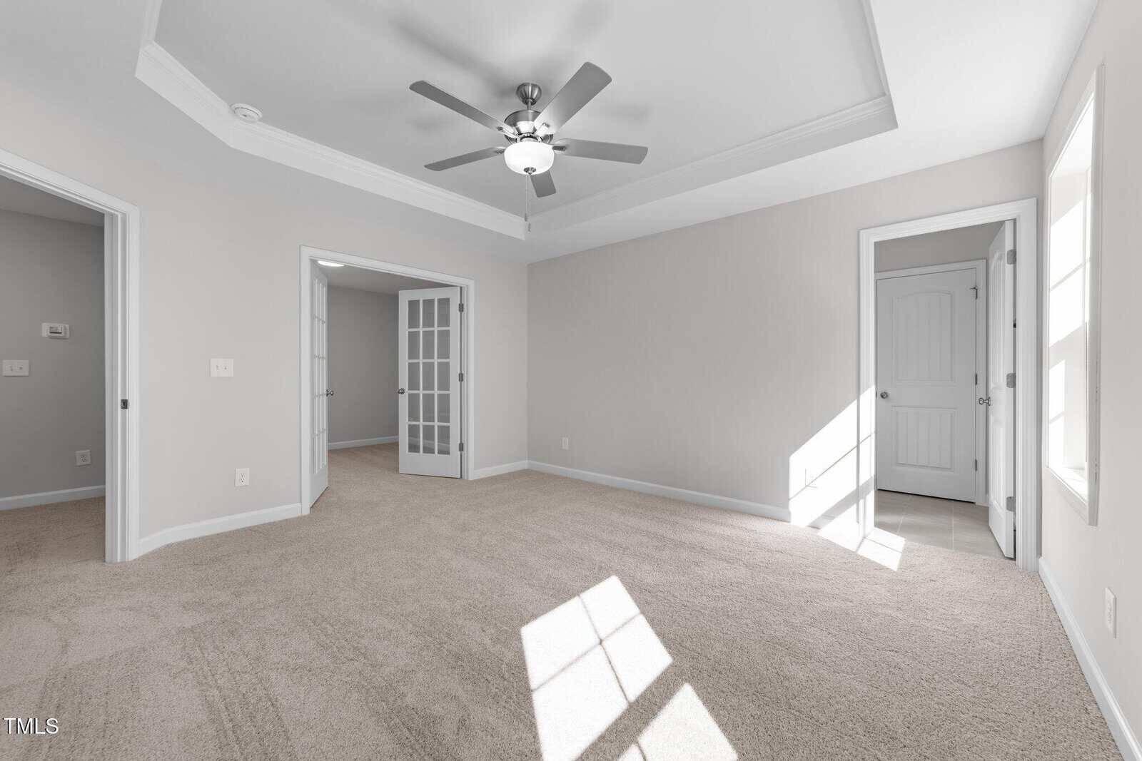 Bright, empty room with beige carpet, gray walls, a ceiling fan, double French doors, a closed white door, and large windows letting in natural light.