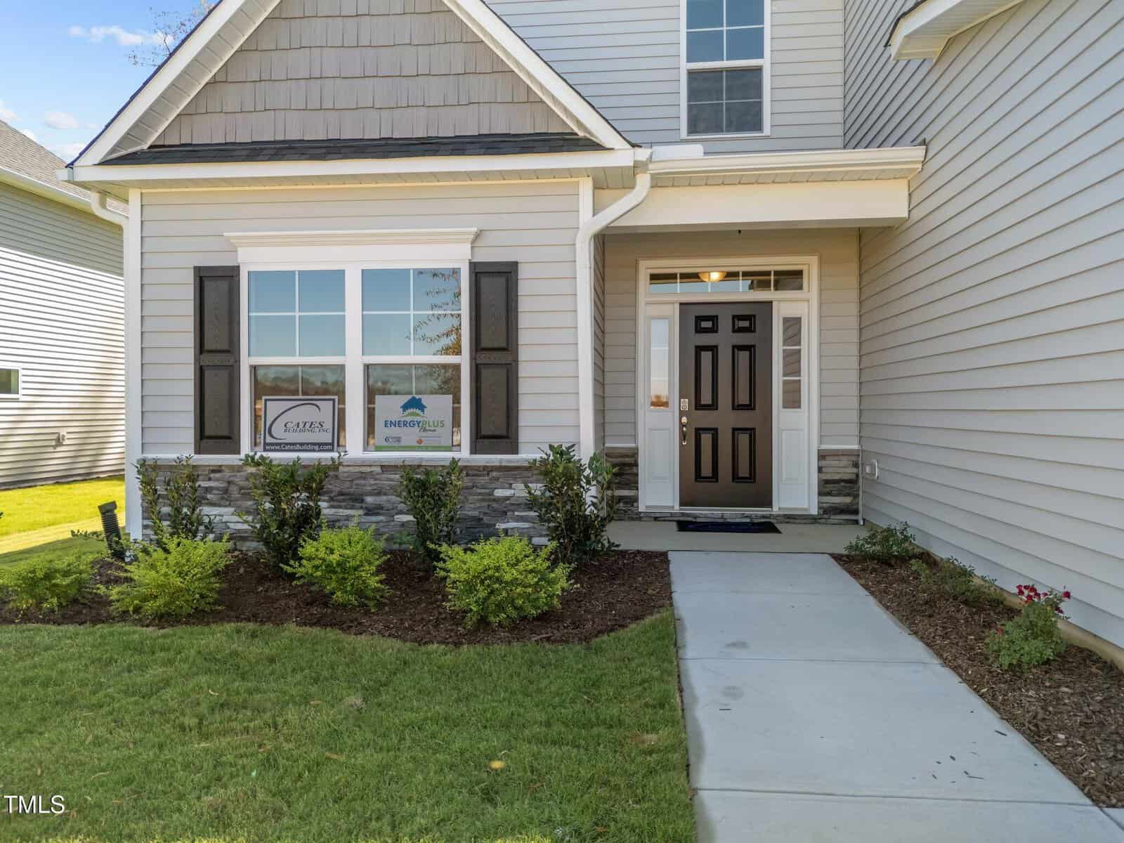 The front entrance of a modern house with light gray siding, stone accent, dark shutters, a black door, small shrubs, and a green lawn bordering a concrete walkway. Signs are displayed in the front window.