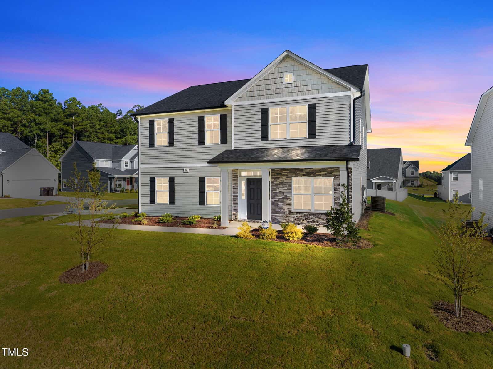 Two-story suburban house with gray siding, black shutters, and a stone accent wall, located at 20 Nebbiolo Drive in Youngsville. The lawn is well-kept, landscaping borders the house, and neighboring homes are visible. The sky is at sunset with shades of pink and purple.