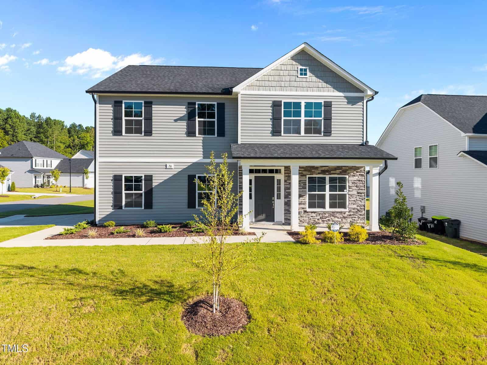 Two-story suburban house with gray siding, black shutters, and partial stone facade. Front porch with white columns, landscaped lawn, young tree, and neighboring homes visible under clear blue sky.